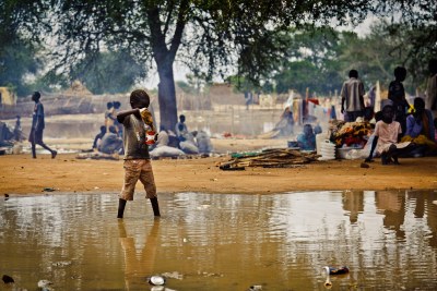 An internally displaced child in Turalei, South Sudan pours water from a mud puddle as others rest under trees.